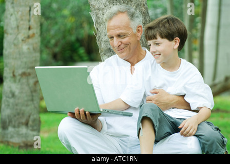 Mature man holding grandson on lap, both looking at laptop computer and smiling Stock Photo