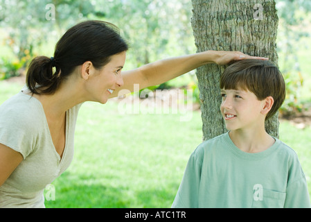 Boy leaning against tree trunk, his mother placing her hand on his head, both smiling Stock Photo