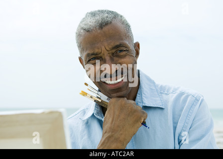 Senior man holding paint brushes outdoors, smiling at camera, canvas in foreground Stock Photo