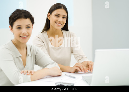 Two businesswomen sitting at desk together next to laptop computer, one smiling at camera Stock Photo