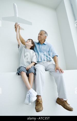 Grandfather and grandson sitting side by side, holding up toy airplane together Stock Photo