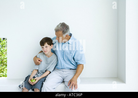 Man sitting with arm around grandson, chatting, both looking away Stock Photo