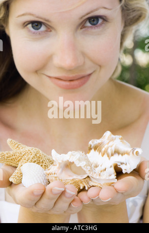 Woman holding up handfuls of seashells, smiling at camera Stock Photo