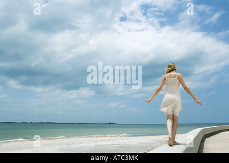 Young woman in sundress walking along low wall at the beach, arms out, rear view Stock Photo