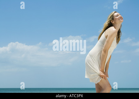 Young woman in sundress standing by the sea, head back, eyes closed, side view Stock Photo