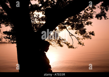 Silhouette of young woman leaning against tree by lake Stock Photo