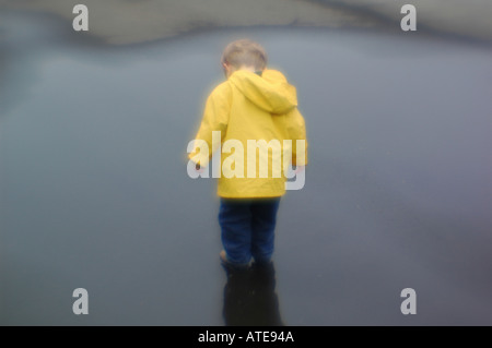 Small wearing yellow rain jacket slicker coat boy playing in puddle of water Stock Photo