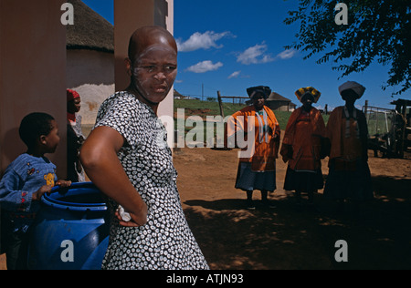 Zulu woman in modern dress daydreaming against pillar, women in traditional attire in background, Uganda Stock Photo