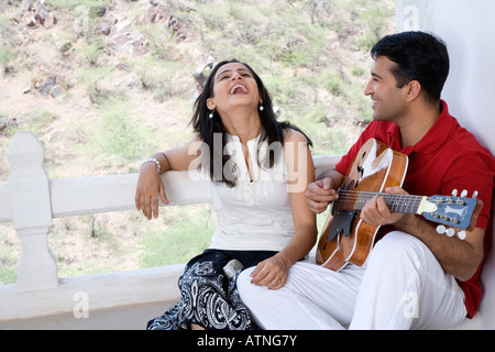 Mid adult man playing a guitar with a mid adult woman sitting beside him and laughing Stock Photo