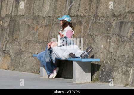 man using cellphone lying on bench with head supported by seated woman Stock Photo