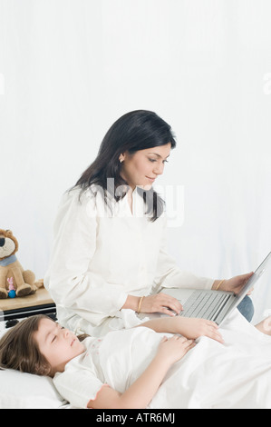 Mid adult woman using a laptop beside her daughter lying on the bed Stock Photo