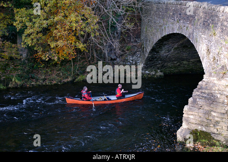 Two people paddling open canoe under a bridge, River Brathay at Clappersgate, near Ambleside, Lake District Nat Park, Cumbria UK Stock Photo