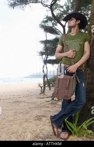Young man carrying a shoulder bag and leaning against a tree trunk on the beach Stock Photo