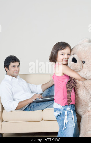 Portrait of a girl hugging a teddy bear with her father using a laptop beside her Stock Photo
