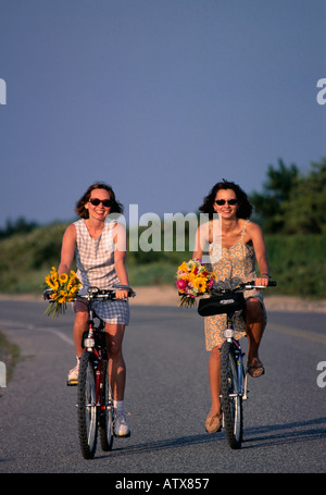 Two young 25 to 30 to 35 years old women sitting on bicycle with basket of flowers, Southampton, Long Island, New York  State, USA Stock Photo