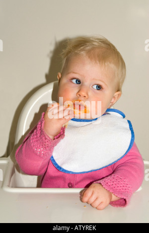 Vertical close up of a pretty Caucasian baby girl sitting at her high chair at dinner time getting in a mess Stock Photo