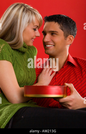 Young enamoured and sitting couple They are both smiling and looking at each other Man is holding box of chocolates Front view Stock Photo
