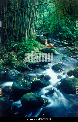 Young woman sitting next to stream in bamboo forest below Akaka Falls on Big Island of Hawaii Stock Photo