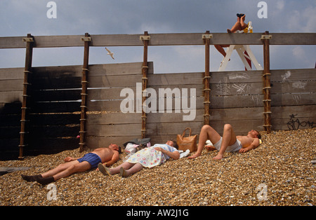 A lifeguard surveys a beach through binoculars while English bathers lie on stone shingle by a coastal groyne. Stock Photo