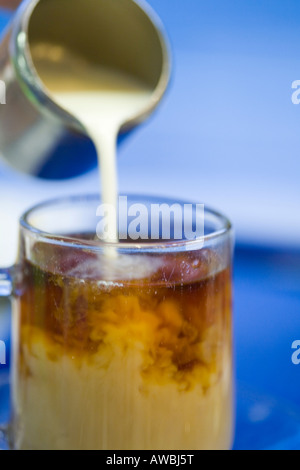 Sequence of pouring milk from a stainless steel jug into a glass of tea against blue background Stock Photo