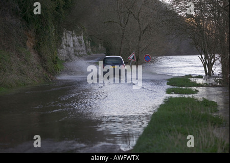 Car being driven through flood water on a road adjacent to a large, fast-flowing and swollen river that has broken its banks.
