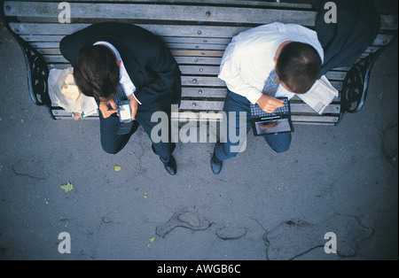 Two business men sitting on a bench using a palm and laptop having lunch Stock Photo
