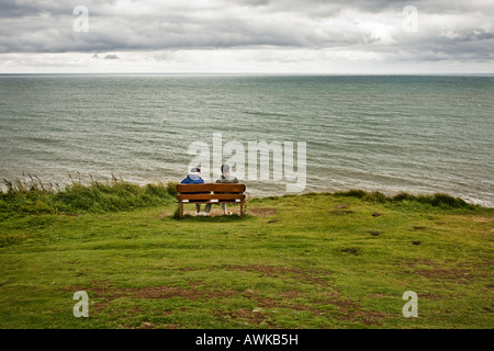 Senior couple sit on wooden bench looking out to sea in autumn / winter, pensioners Stock Photo