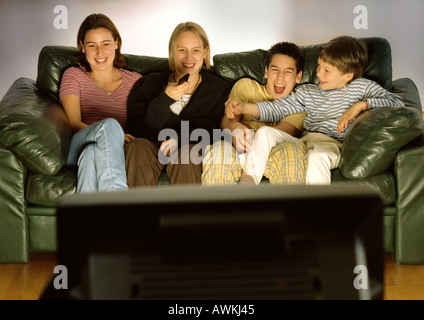 Three young people and one child sitting on green couch together, laughing, blurred rear view of tv in foreground Stock Photo