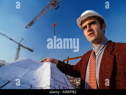 Man wearing hard hat, holding blueprint, cranes in background, low angle view Stock Photo