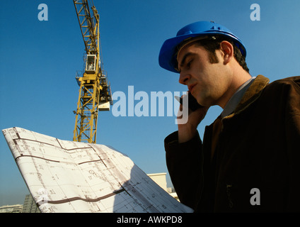 Man wearing hard hat, holding cell phone and blueprints Stock Photo