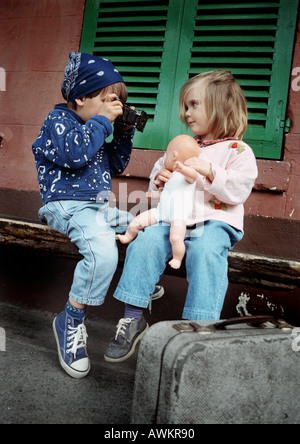 Two girls sitting on bench, one holding camera, suitcase on ground Stock Photo