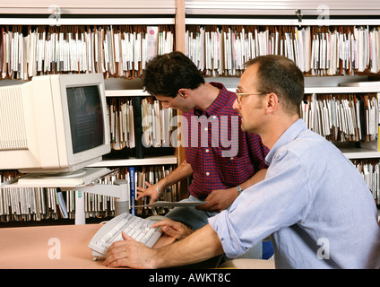 Man using computer, other man looking at files on shelves Stock Photo