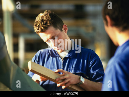 Man in blue coveralls holding plank of wood, second man rear view, blurred in foreground Stock Photo