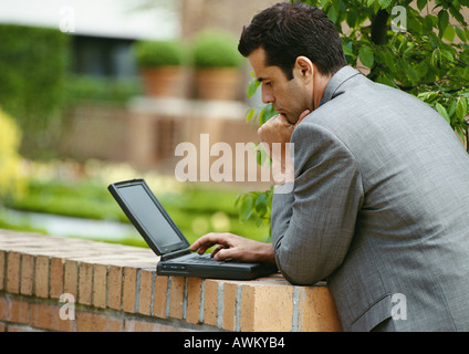 Businessman using laptop computer outside, leaning against low brick wall, side view Stock Photo