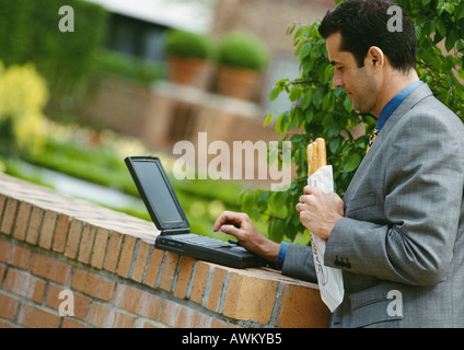 Businessman using laptop computer while eating sandwich outside Stock Photo