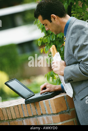 Businessman using laptop computer outside, holding sandwich Stock Photo