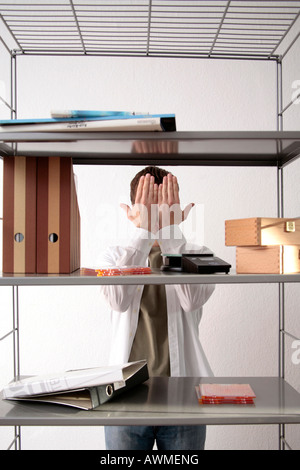 Young man standing behind a bookshelf, covering his eyes with his hands Stock Photo