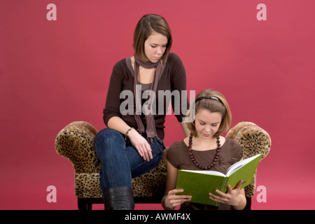 Two girls, pre-teens, early teens sitting on a tiger-print couch reading a book Stock Photo