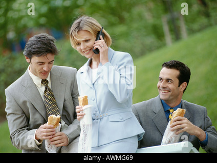 Three business people holding sandwiches outdoors, one using cell phone Stock Photo