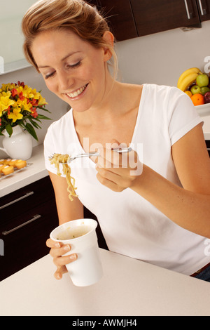 WOMAN IN KITCHEN EATING POT NOODLE Stock Photo