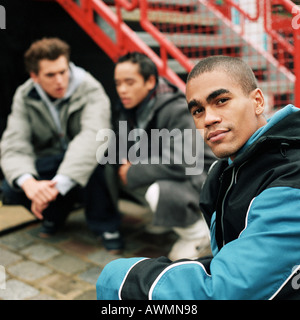 Focus on young man looking into camera in foreground, two young men squatting in background Stock Photo