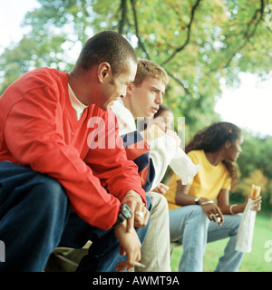 Young people sitting together on bench outside, some with sandwiches Stock Photo