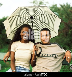 Young man and young woman smiling and sitting under umbrella outside Stock Photo