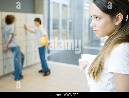 Teenage girl holding notebook, looking away, two teen boys standing near lockers in background Stock Photo