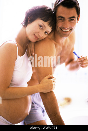 Man brushing teeth in bathroom, pregnant woman leaning on his shoulder Stock Photo