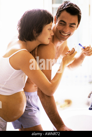 Man brushing teeth in bathroom, pregnant woman leaning on his shoulder Stock Photo