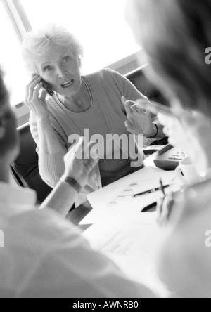 Mature businesswoman in conference, using cell phone, other businesspeople blurred in foreground, B&W Stock Photo