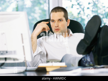 Businessman holding glasses next to face, looking at computer screen, sitting with feet on desk Stock Photo