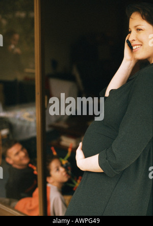 Pregnant woman standing, using cell phone and holding stomach, man and child sitting in background Stock Photo