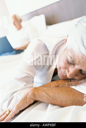 Senior woman sleeping on bed, man using laptop in background Stock Photo